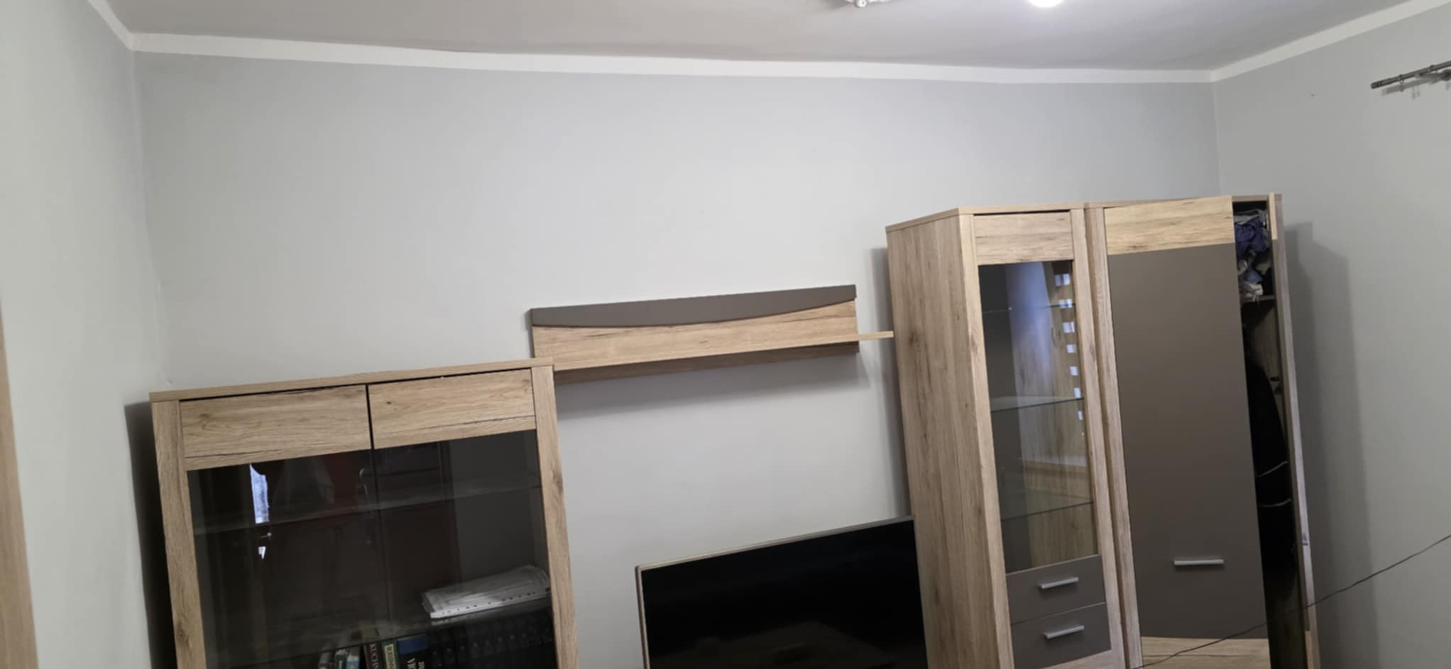 Jasnoszary salon po remoncie: ściany pomalowane na gładko, widoczna drewniana meblościanka z szarymi akcentami. Minimalistyczny styl.