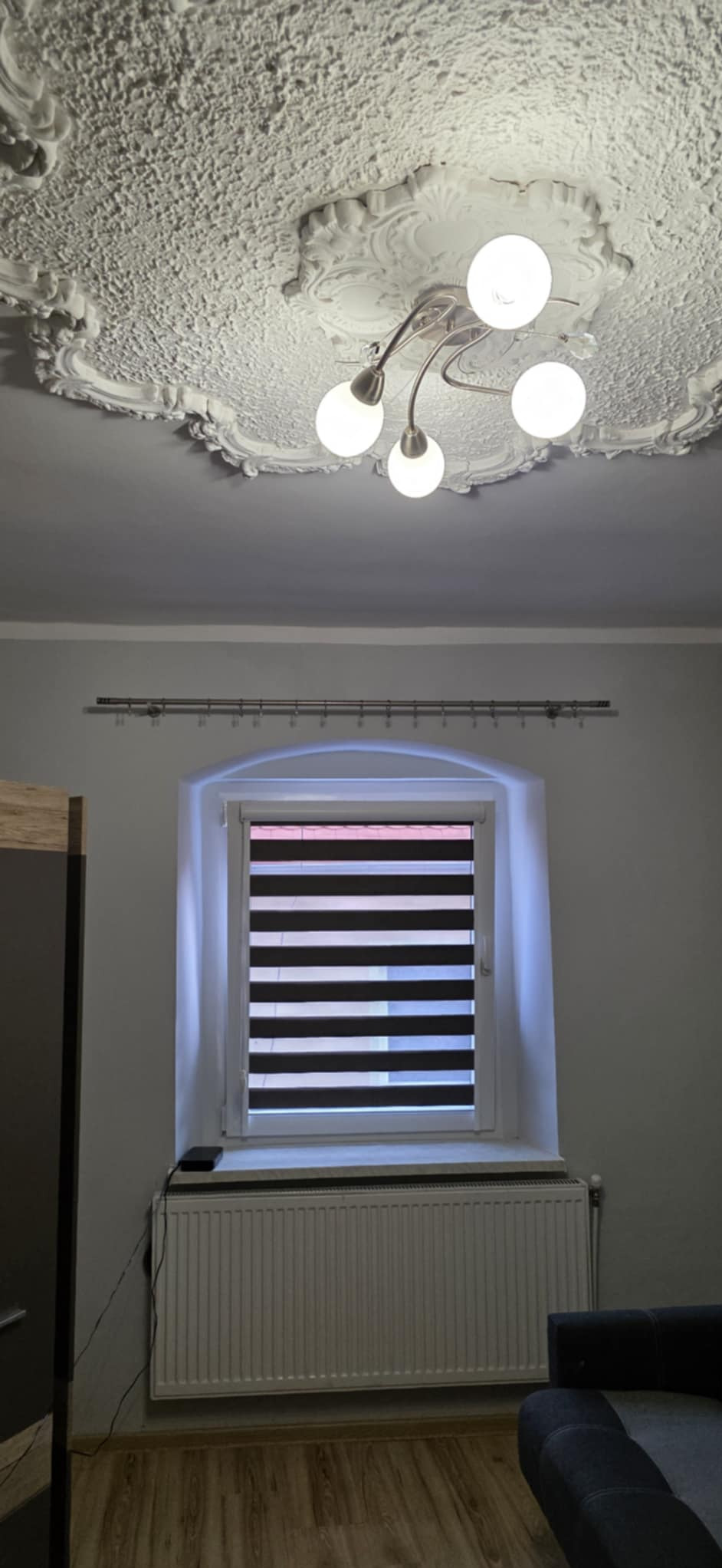 Pokój z dekoracyjnym sufitem sztukatorskim i nowoczesnym oświetleniem, okno z roletami dzień-noc, grzejnik i fragment mebla. Ujęcie z dołu.