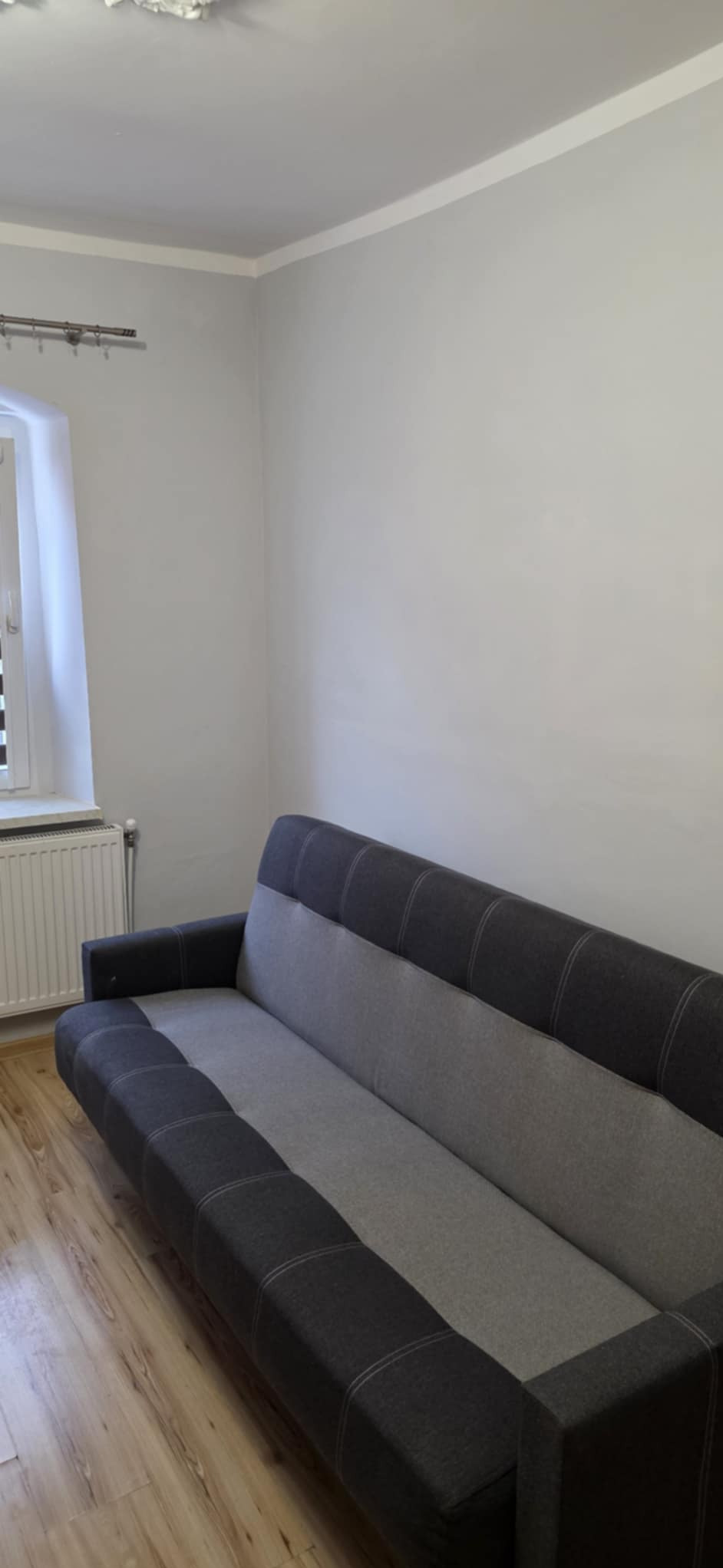 Jasnoszary pokój po malowaniu z szarą sofą. Widoczne gładkie ściany, okno i drewniana podłoga. Minimalistyczny wystrój po remoncie.