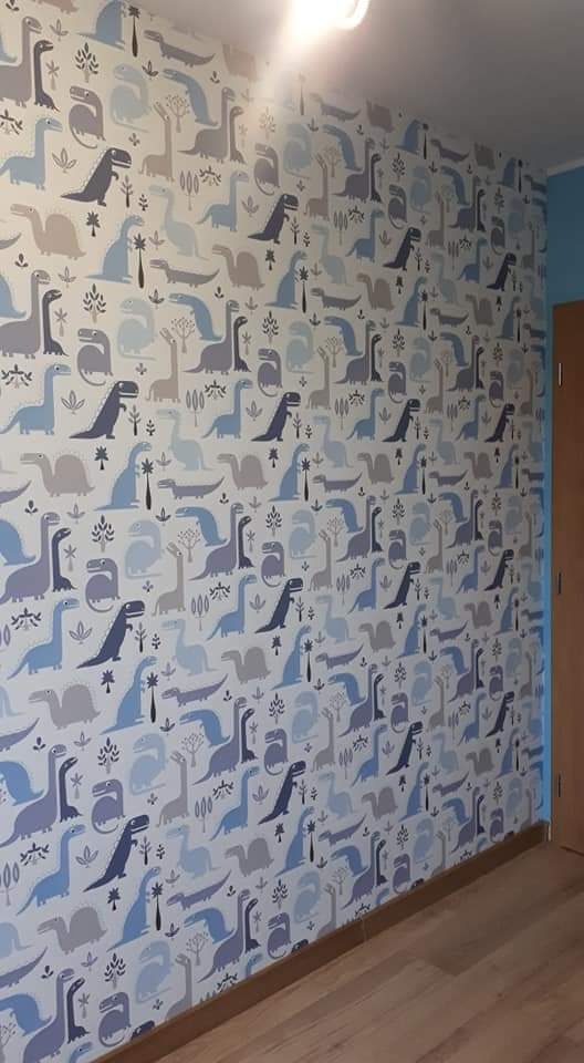 Ściana w pokoju dziecięcym pokryta tapetą z motywem dinozaurów w odcieniach szarości i błękitu, widoczna podłoga z paneli.