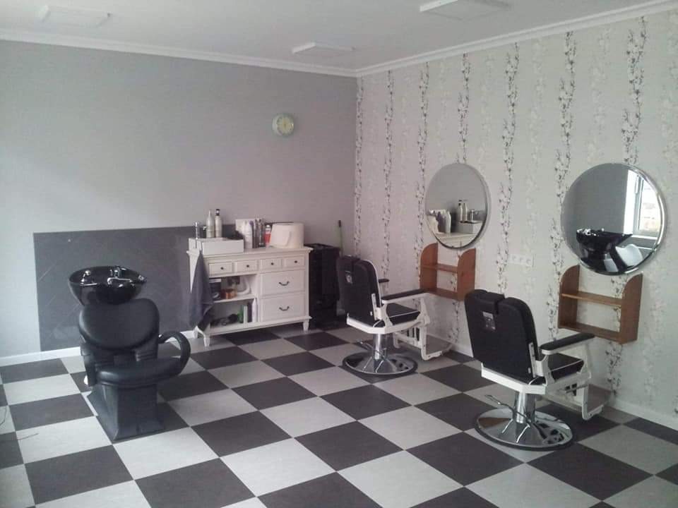 Umeblowane wnętrze salonu fryzjerskiego z dwoma fotelami, umywalką fryzjerską, komodą z kosmetykami i lustrami na ścianie z tapetą w motywy roślinne.