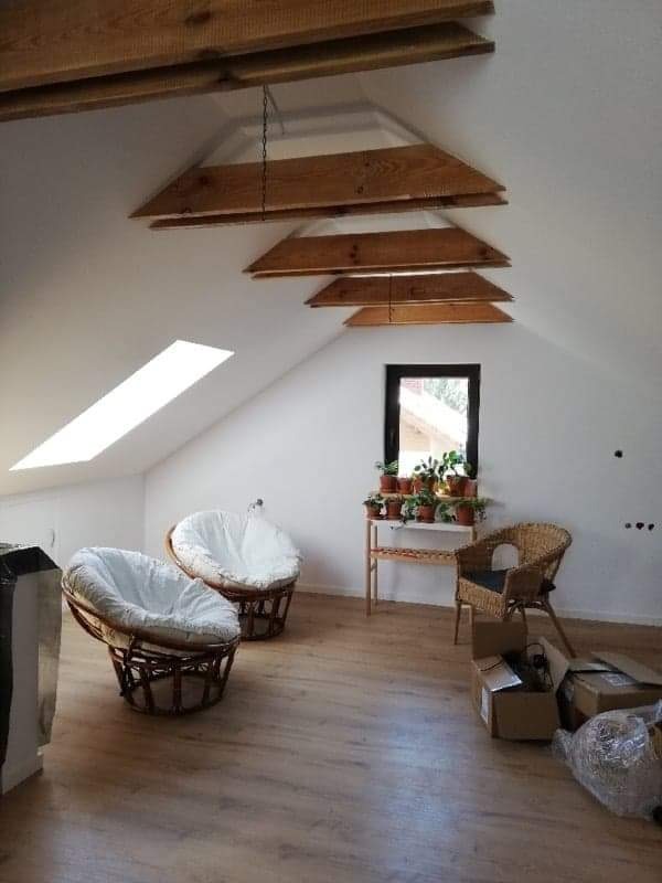 Poddasze po remoncie z drewnianymi belkami dekoracyjnymi, dwoma fotelami papasan, drewnianą podłogą i oknem z roślinami, częściowo jeszcze w kartonach po przeprowadzce.