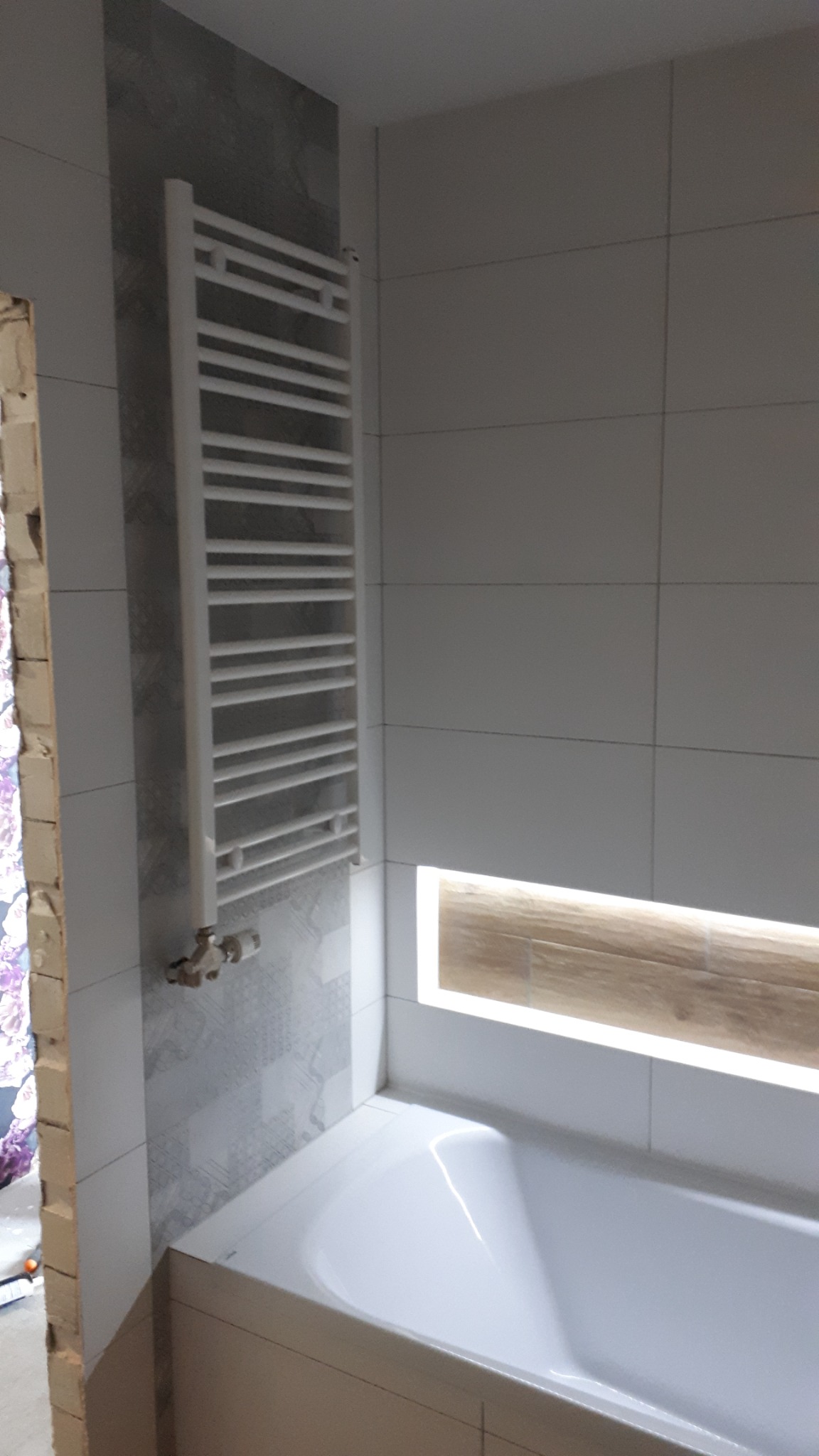 Biała drabinka grzejnikowa zamontowana na ścianie z szarymi płytkami w łazience, widoczna wanna i podświetlona wnęka z drewnopodobnym wykończeniem.