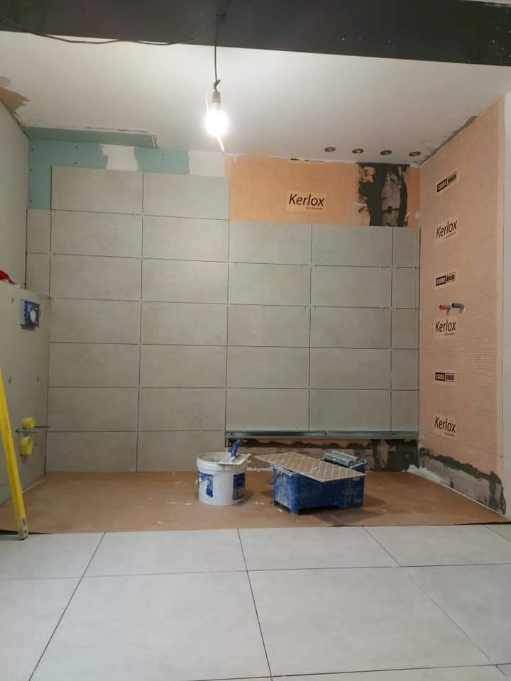 Układanie płytek w łazience w trakcie prac: częściowo wyłożone ściany, widoczne narzędzia i materiały budowlane, pomarańczowa hydroizolacja Kerlox na ścianie.