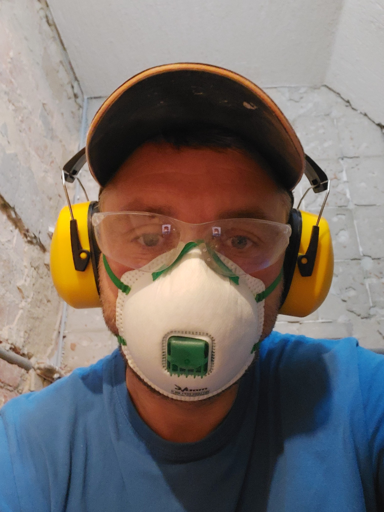 Pracownik w masce ochronnej, okularach i słuchawkach na tle ściany w trakcie przygotowań do prac remontowych.