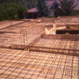 Zbrojenie stropu budynku jednorodzinnego z widocznymi deskami szalunkowymi i wystającymi prętami zbrojeniowymi; w tle dom z czerwonym dachem i zielona roślinność.