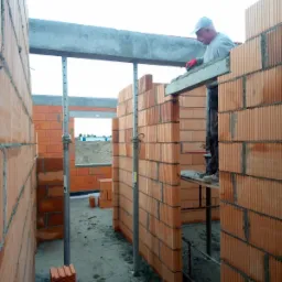 Wznoszenie ścian parteru z bloczków ceramicznych Porotherm, widoczny pracownik na rusztowaniu, podpory stropowe i fragment widoku na okolicę przez otwór okienny.