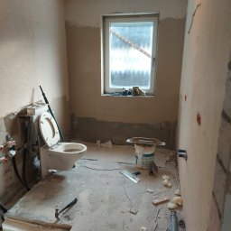 Top Glazur - Łazienka w trakcie remontu: surowe ściany, zamontowana toaleta z otwartą klapą, widoczne narzędzia i materiały budowlane, okno z częściowo zasłoniętą szybą.