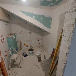 Top Glazur - Łazienka w trakcie układania białych płytek z użyciem czerwonych krzyżyków dystansowych, widoczna drabina, poziomica, wiadro z klejem i częściowo wykończony sufit pod skosem.
