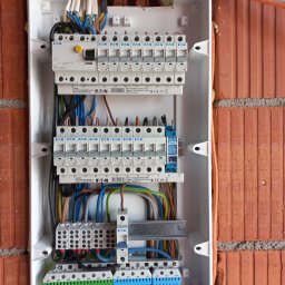 Top Glazur - Otwarta skrzynka elektryczna z zainstalowanymi bezpiecznikami i podłączonymi kablami, zamontowana na ścianie z cegły. Widoczne elementy marki Eaton oraz Eltako.