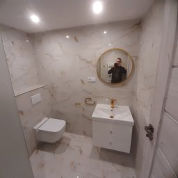 Top Glazur - Wykończona łazienka z marmurowymi płytkami, okrągłym lustrem z odbiciem fotografa, białą szafką z umywalką i złotą armaturą oraz wiszącą toaletą.