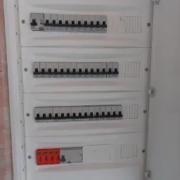 Otwarta skrzynka elektryczna z czterema rzędami bezpieczników i zabezpieczeniem przeciwprzepięciowym SIMTEC na dole, widoczne przewody u dołu.