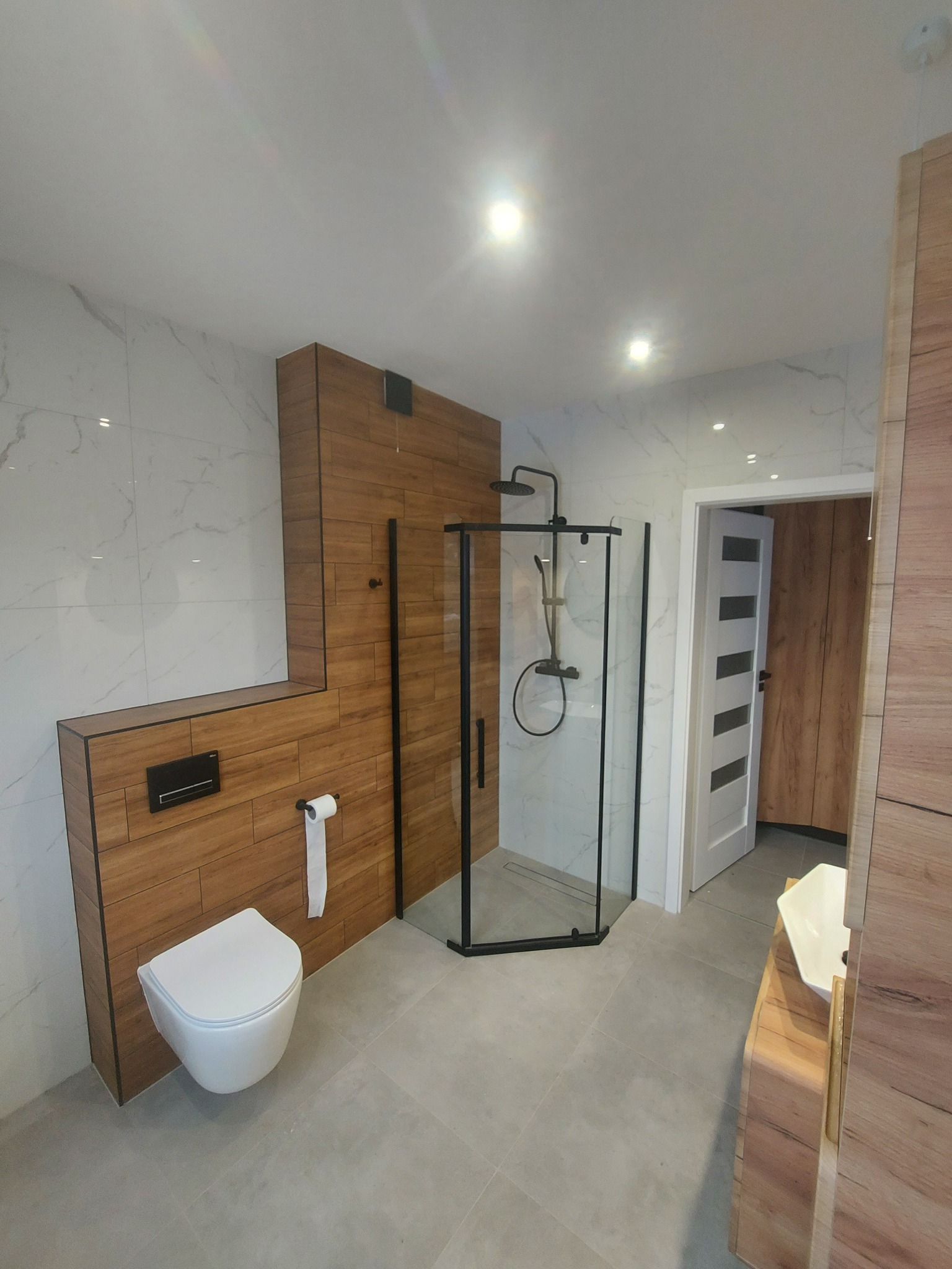 Nowoczesna łazienka z kabiną prysznicową typu walk-in z czarnymi profilami, podwieszaną toaletą i umywalką nablatową na drewnianej szafce, wykończona płytkami imitującymi drewno i marmur.