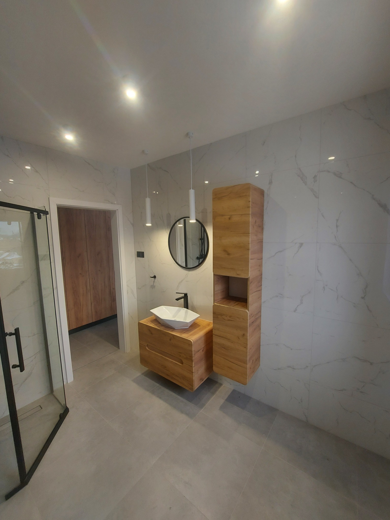 Nowoczesna łazienka z drewnianą szafką pod umywalką, wiszącą szafką, okrągłym lustrem i kabiną prysznicową z czarnymi profilami, wykończona płytkami imitującymi marmur.