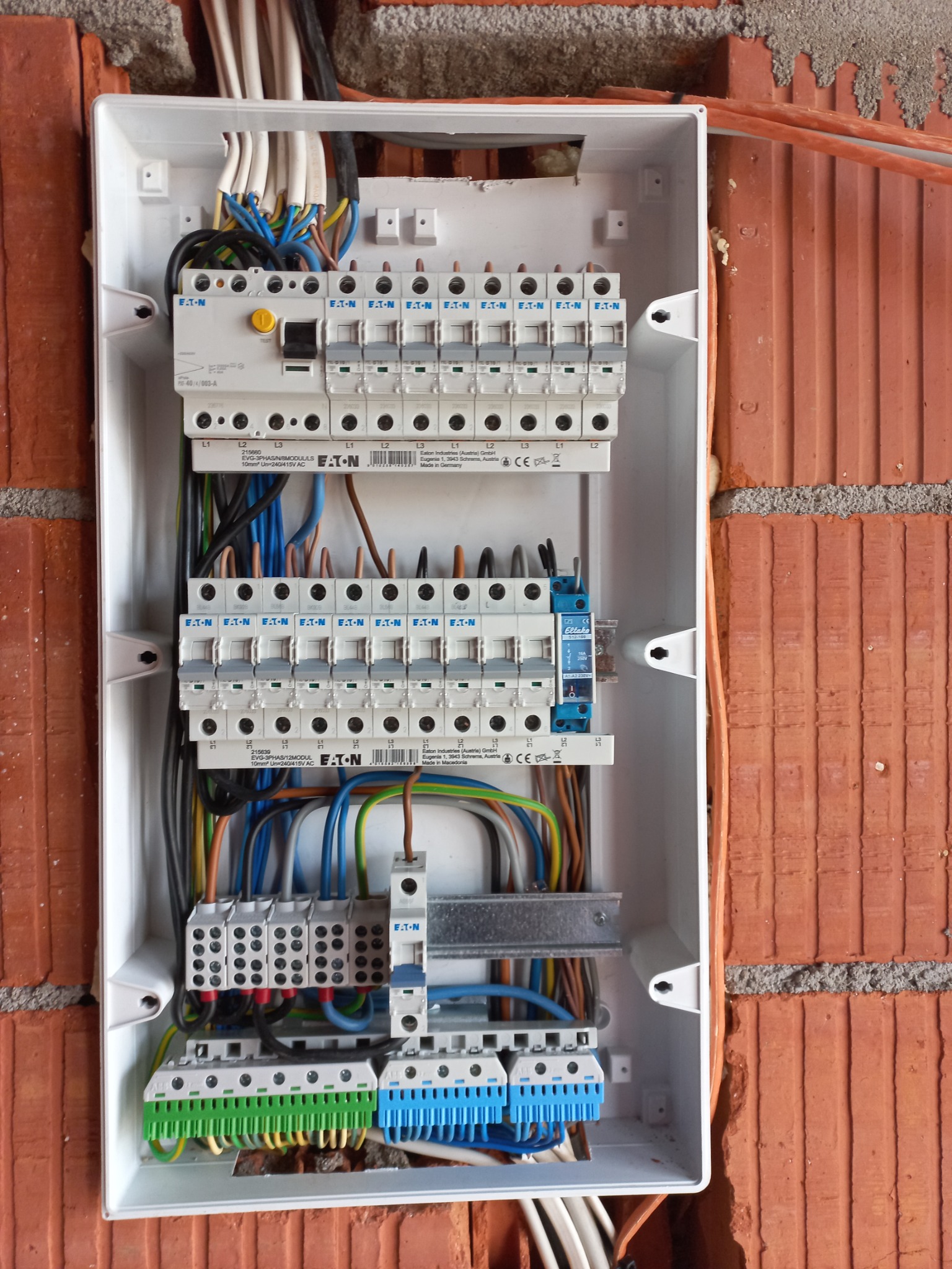 Otwarta skrzynka elektryczna z zainstalowanymi bezpiecznikami i podłączonymi kablami, zamontowana na ścianie z cegły. Widoczne elementy marki Eaton oraz Eltako.