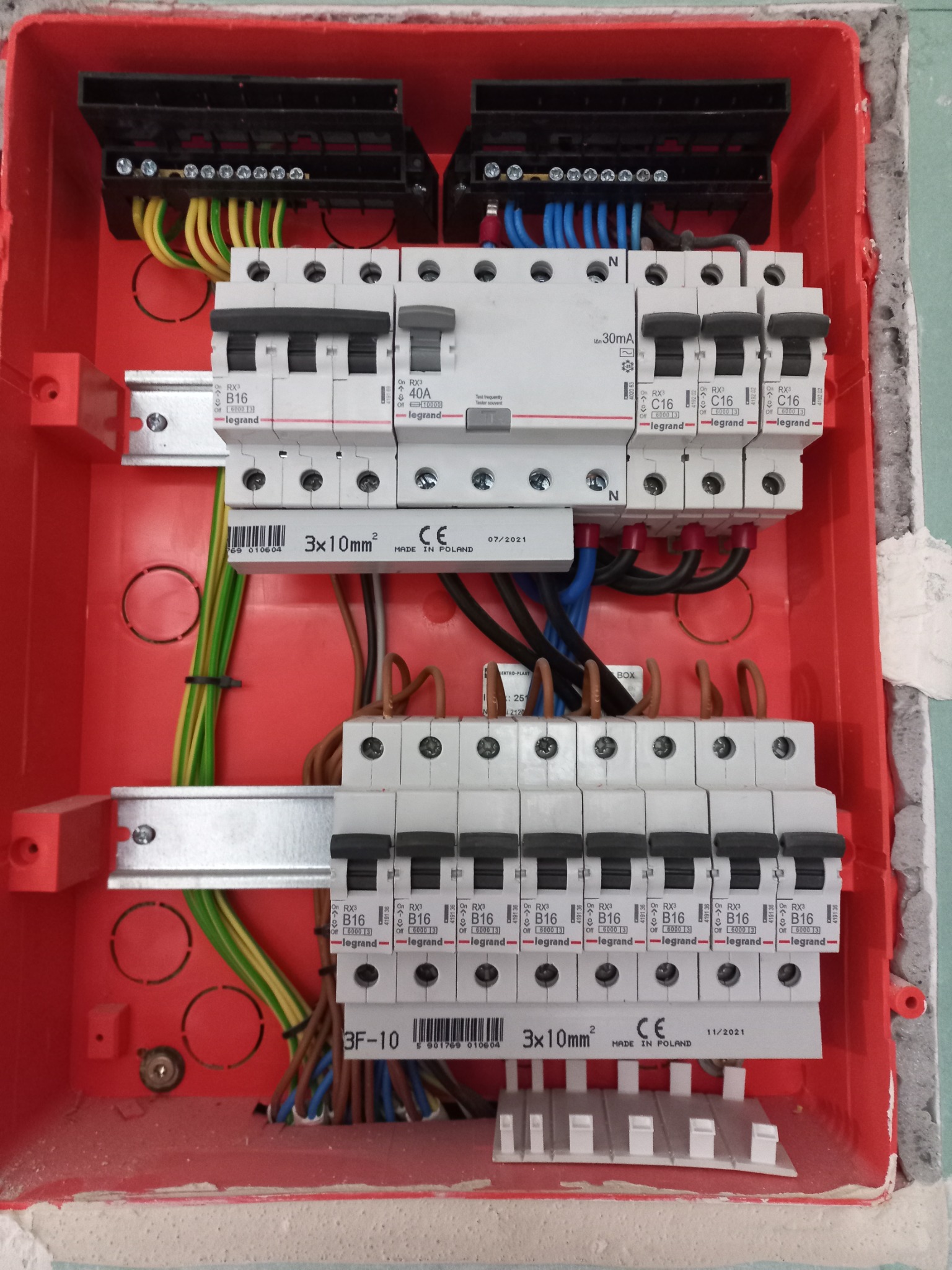 Rozdzielnia elektryczna w czerwonej obudowie, widoczne bezpieczniki Legrand B16 i C16, przewody zasilające w różnych kolorach, montaż w trakcie prac wykończeniowych.