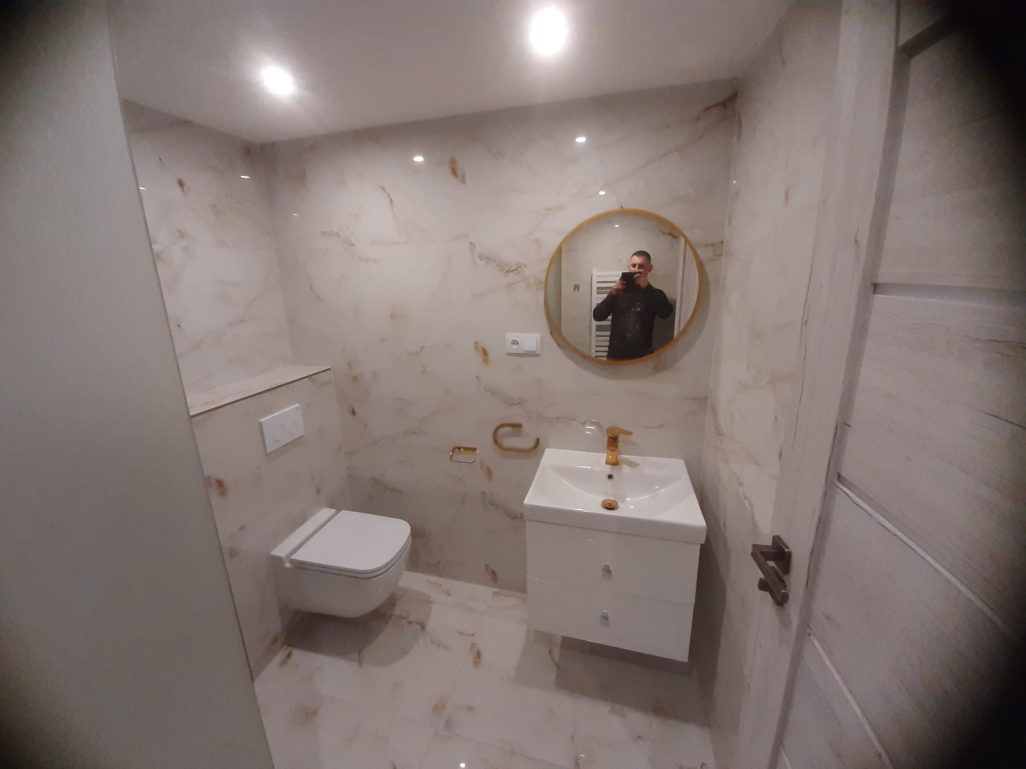 Wykończona łazienka z marmurowymi płytkami, okrągłym lustrem z odbiciem fotografa, białą szafką z umywalką i złotą armaturą oraz wiszącą toaletą.