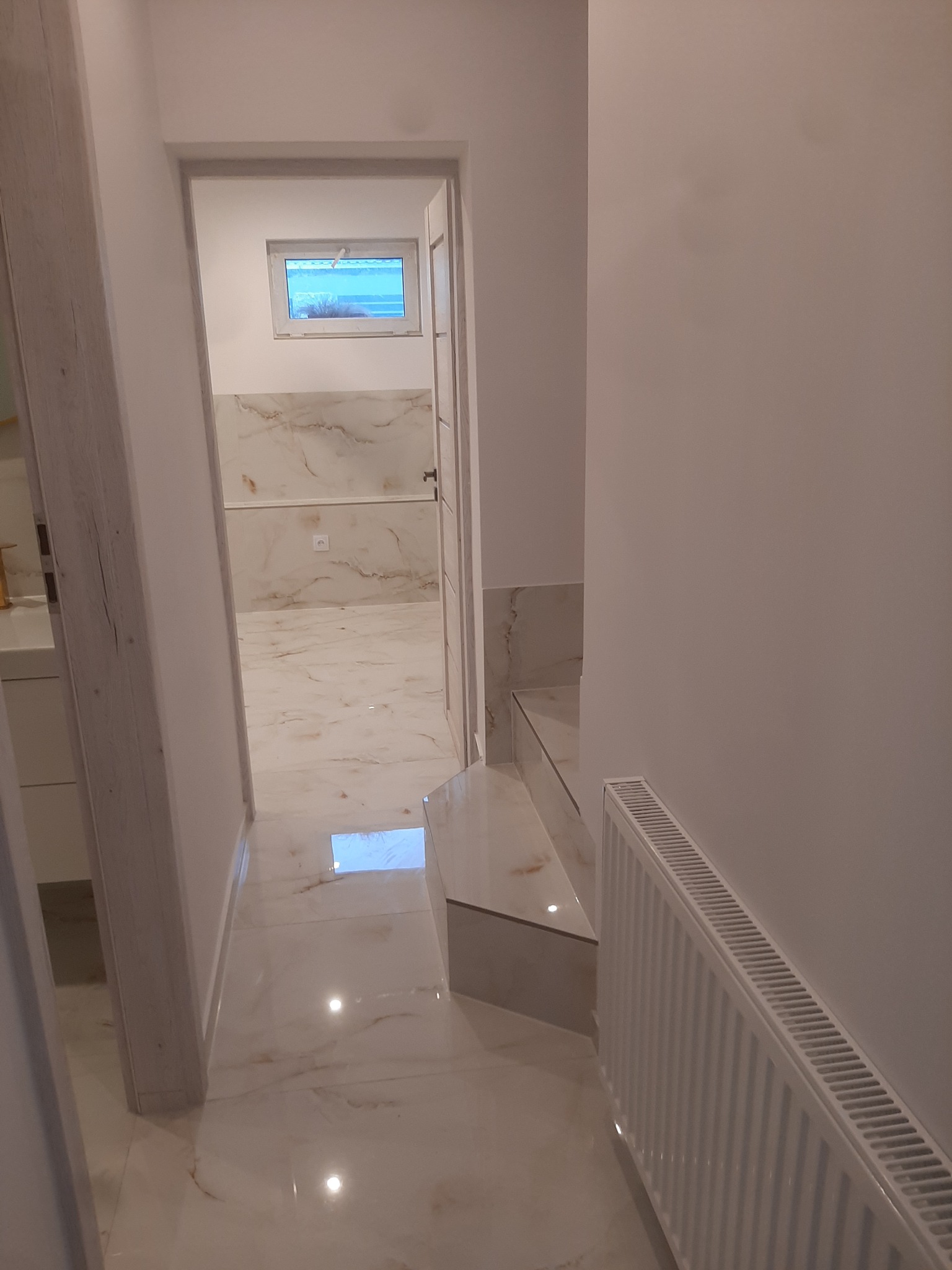 Wykończony korytarz z marmurową podłogą i grzejnikiem żeberkowym, widok przez otwarte drzwi na łazienkę z podobnym wykończeniem.