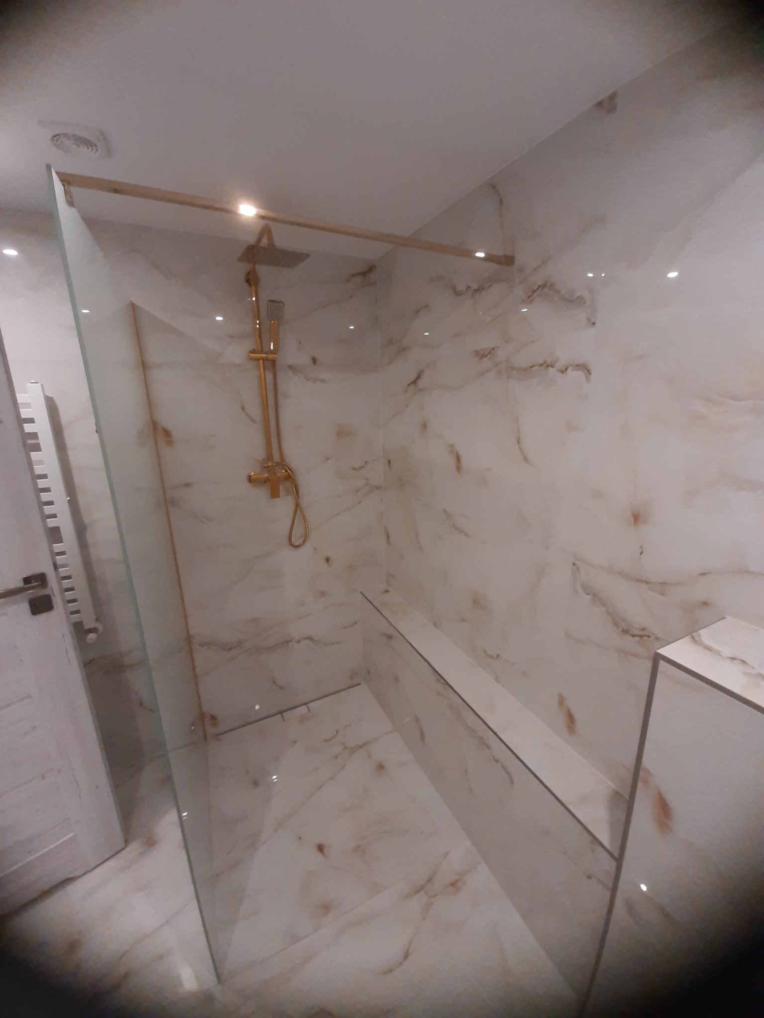 Nowoczesna kabina prysznicowa typu walk-in z marmurowymi płytkami i złotą armaturą, widoczny grzejnik drabinkowy i zabudowana półka.