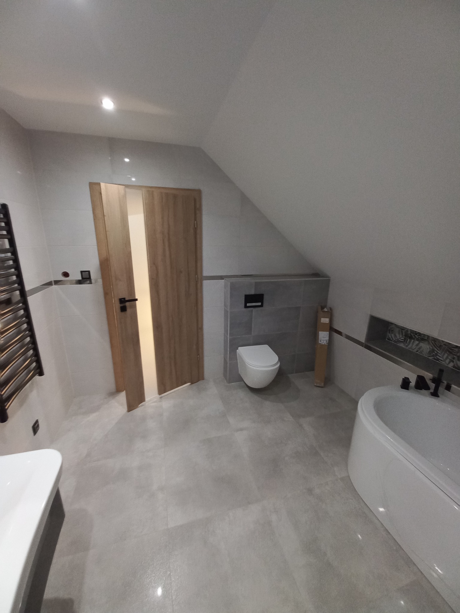 Nowoczesna łazienka na poddaszu z szarymi płytkami, białą armaturą i drewnianymi drzwiami z przeszkleniem, widoczny grzejnik drabinkowy.