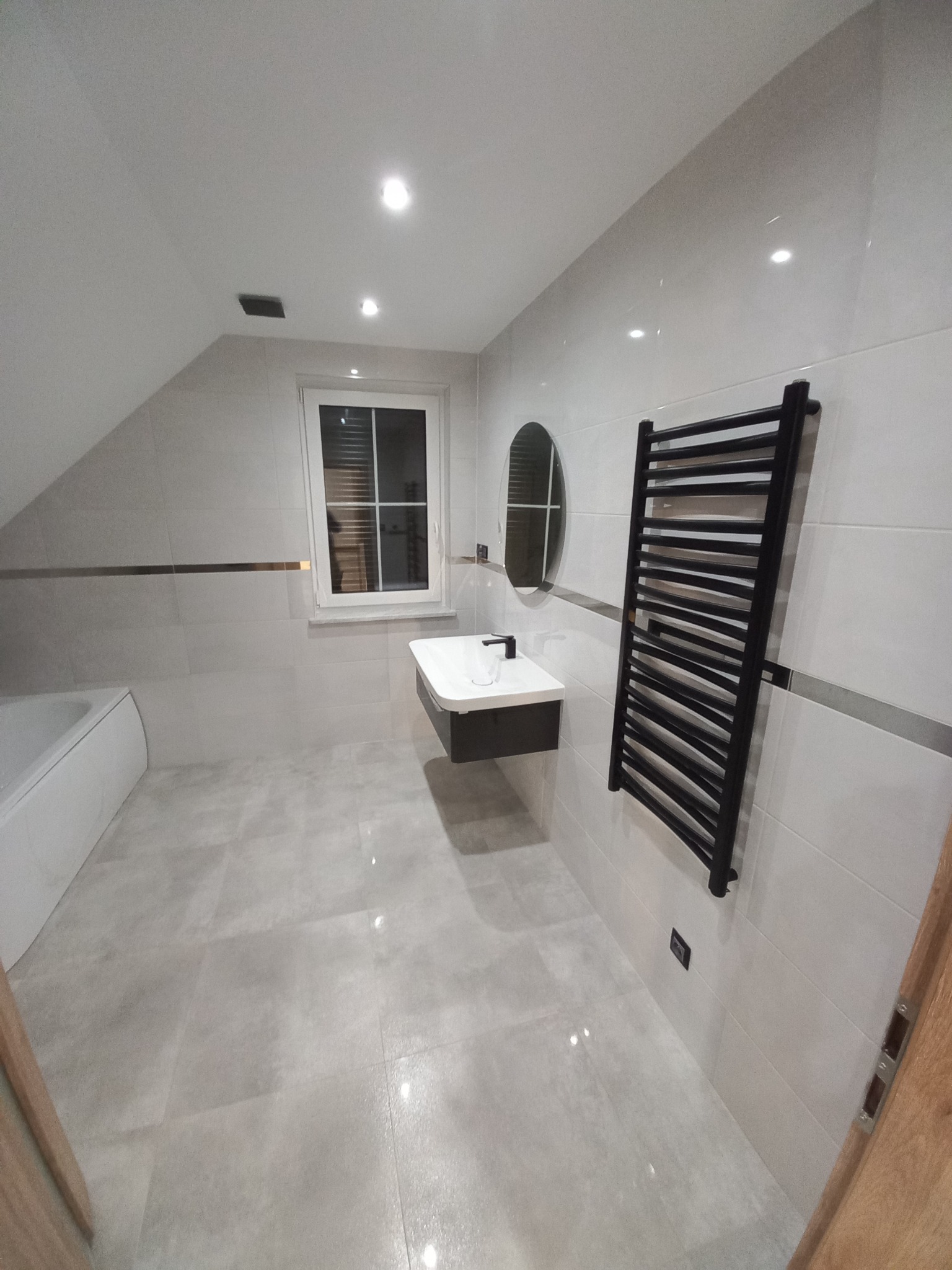 Nowoczesna łazienka na poddaszu z białymi płytkami, czarnym grzejnikiem drabinkowym i wiszącą szafką z umywalką, oświetlona punktowo.