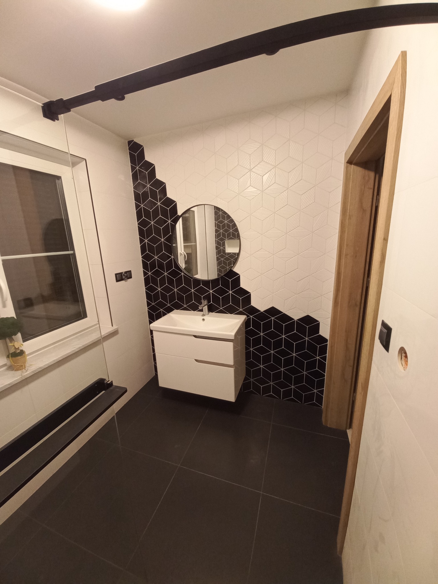 Nowoczesna łazienka z czarno-białymi geometrycznymi płytkami, okrągłym lustrem nad białą szafką umywalkową i ciemną podłogą z płytek, widoczna drewniana ościeżnica drzwi.
