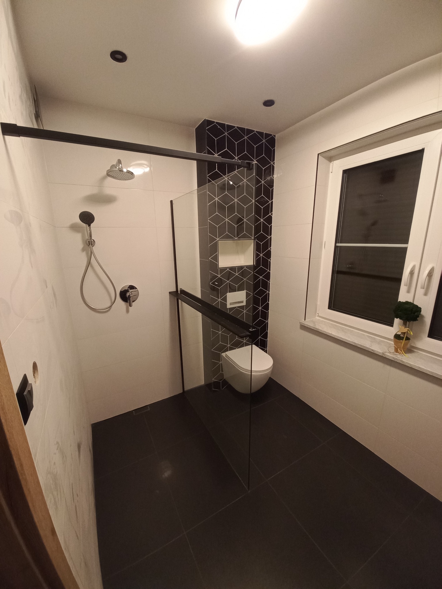 Nowoczesna łazienka z kabiną prysznicową typu walk-in z czarnymi elementami, białą armaturą i czarno-białymi geometrycznymi płytkami na ścianie.