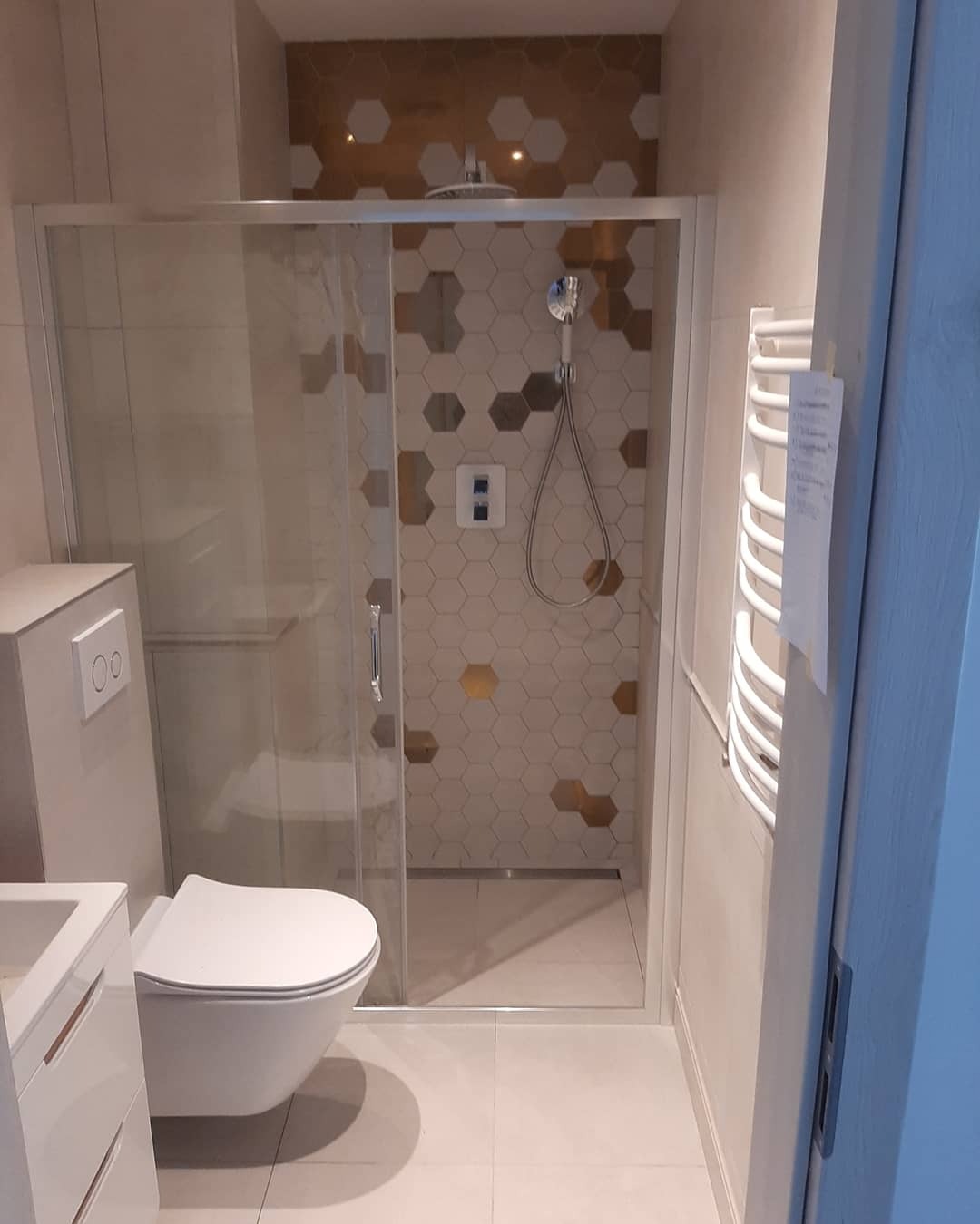 Nowoczesna łazienka z kabiną prysznicową typu walk-in, wykończona heksagonalnymi płytkami w odcieniach bieli i złota, z wiszącą toaletą i grzejnikiem drabinkowym.