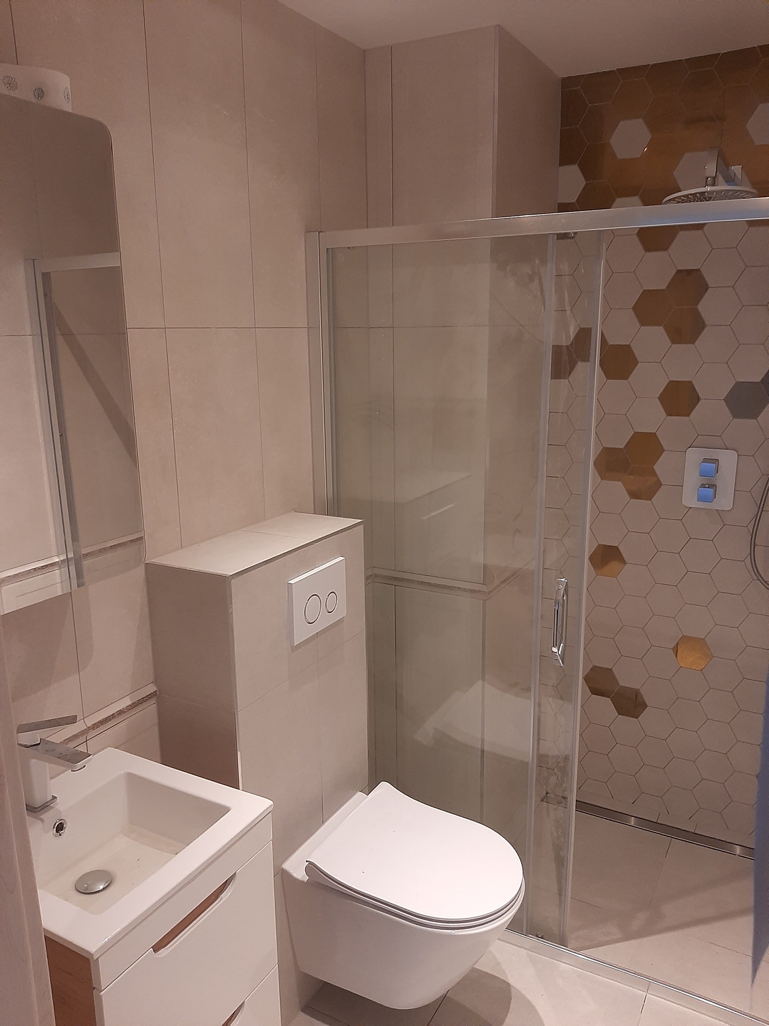 Nowoczesna łazienka w beżach z kabiną prysznicową wykończoną heksagonalnymi płytkami w odcieniach bieli i złota, wisząca toaleta i umywalka z szafką.