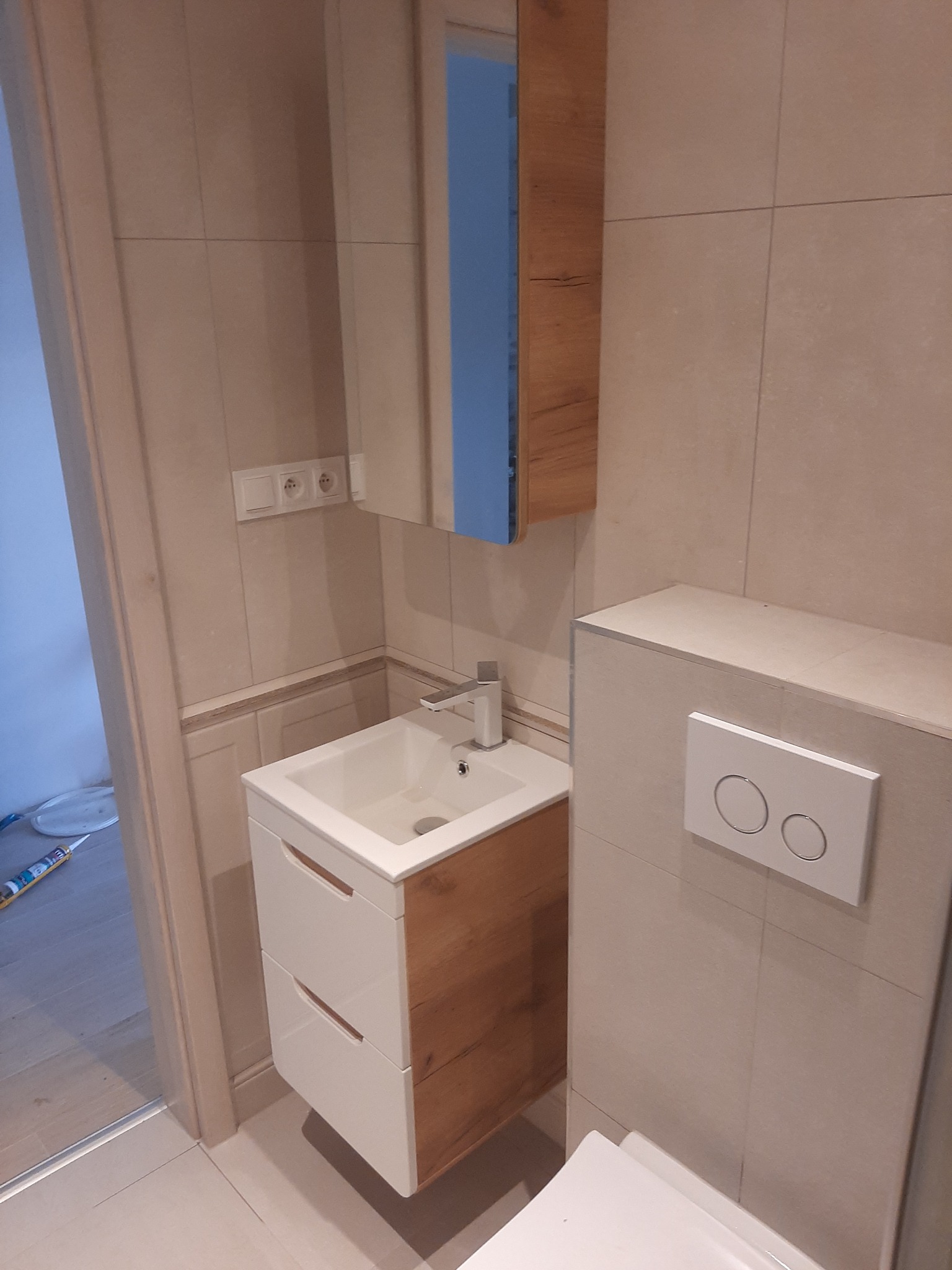 Nowoczesna łazienka z beżowymi płytkami, wiszącą szafką z umywalką z drewnianymi frontami i lustrem, oraz podwieszanym WC z przyciskiem spłukującym