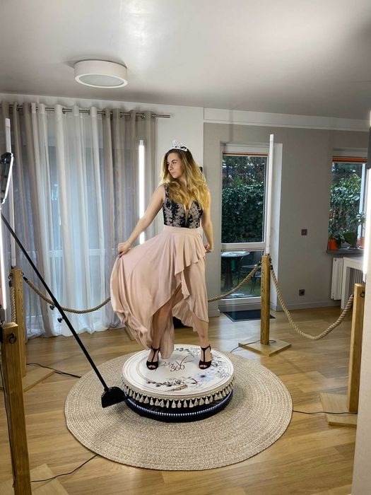 Kobieta w koronie i sukni pozuje na obrotowej platformie otoczonej linami i oświetleniem LED w pomieszczeniu z oknami.