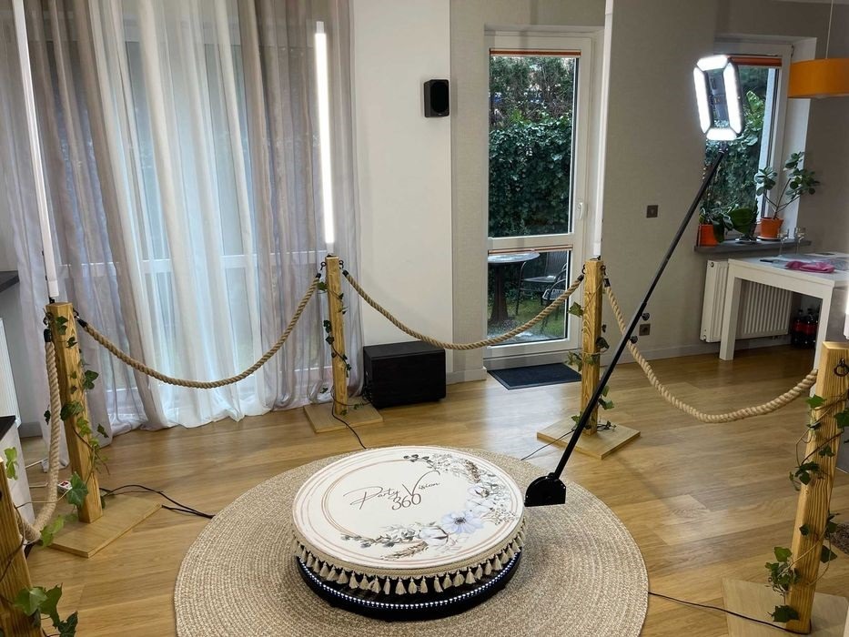 Okrągła platforma obrotowa z dekoracjami kwiatowymi i napisem 'Party Vision 360', otoczona linami z drewnianymi słupkami i sztucznym bluszczem, oświetlona lampami LED, na tle okna z widokiem na ogród.