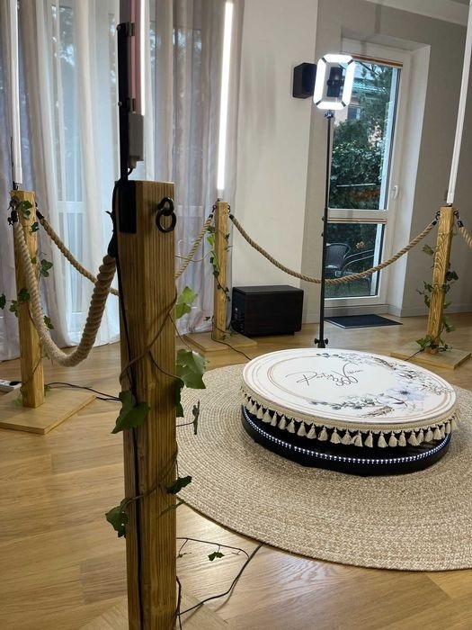 Okrągła fotobudka 360 z dekoracją kwiatową i oświetleniem LED, otoczona drewnianymi słupkami z linami i sztucznym bluszczem, ustawiona na beżowym dywanie.