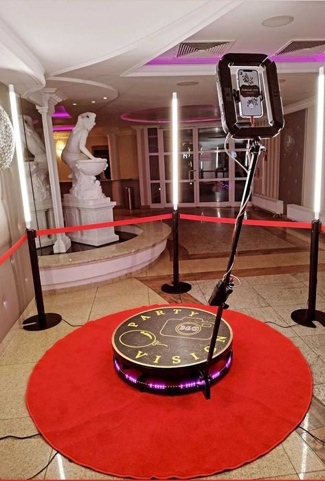 Oświetlona platforma obrotowa z uchwytem na telefon, otoczona czerwonym dywanem i słupkami z taśmą, w eleganckim wnętrzu z rzeźbami.