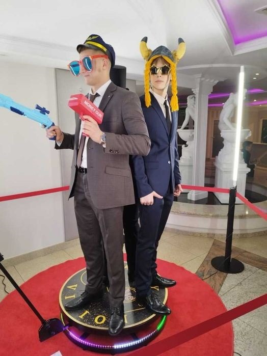 Dwaj mężczyźni w garniturach i zabawnych okularach i kapeluszach pozują z plastikowymi pistoletami na tle wnętrza z kolumnami i statuami, stojąc na podświetlanej platformie.