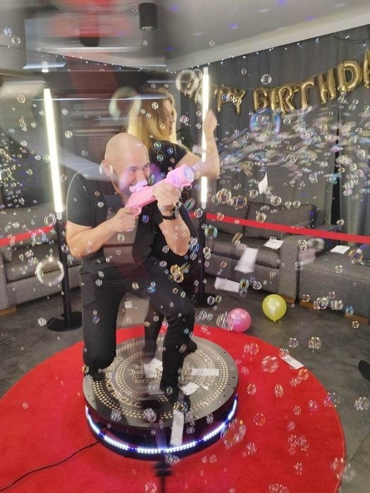 Uczestnicy imprezy na platformie obrotowej, mężczyzna strzela bańkami mydlanymi z różowego pistoletu, w tle dekoracja urodzinowa.