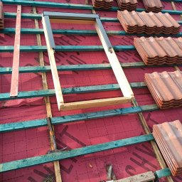 Usługi dekarskie Niepoczołowice - Montaż okna dachowego w konstrukcji dachu z czerwonej membrany i ułożonych dachówek karpiówek w stosach, widoczne drewniane łaty i fragmenty narzędzi.