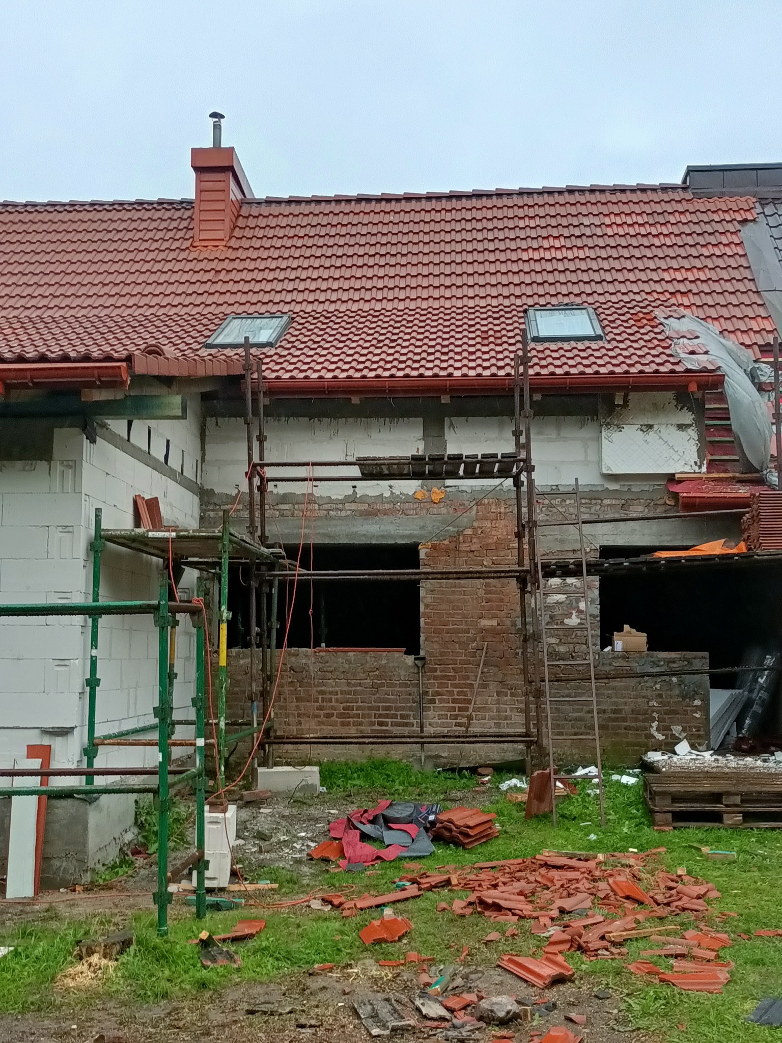 Budynek w trakcie remontu dachu z widocznymi rusztowaniami, częściowo położoną czerwoną dachówką, oknami dachowymi i stertą potłuczonych dachówek na ziemi.