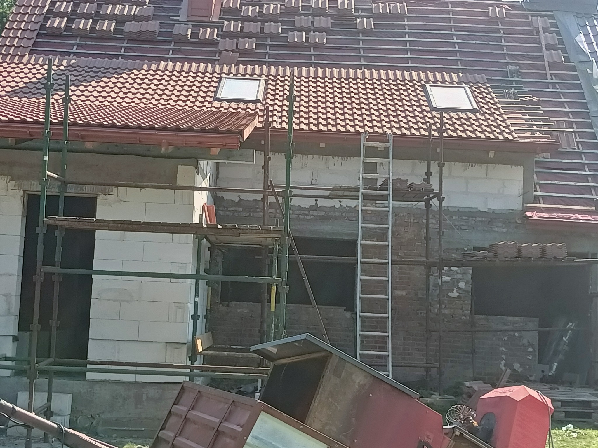 Budowa domu jednorodzinnego w trakcie kładzenia dachówki, widoczne rusztowania, okna dachowe i częściowo ułożone pokrycie z dachówki ceramicznej.