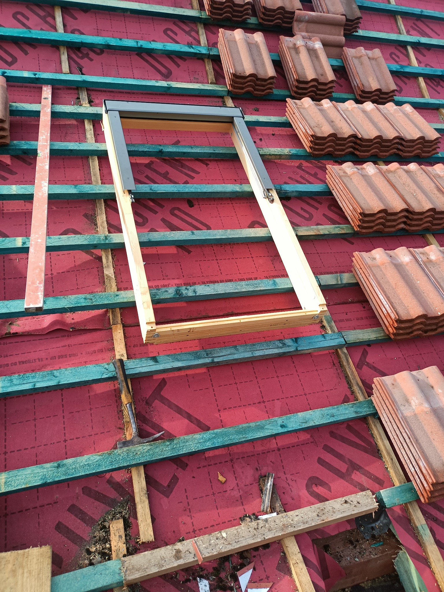 Montaż okna dachowego w konstrukcji dachu z czerwonej membrany i ułożonych dachówek karpiówek w stosach, widoczne drewniane łaty i fragmenty narzędzi.