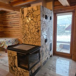 PEKASA Serhii Lypianchyk - Rustykalny kominek obudowany mozaiką z kamienia w drewnianym wnętrzu. Widoczne palenisko z szybą i fragment drewnianej ściany z krzyżem. Za oknem zimowy krajobraz.