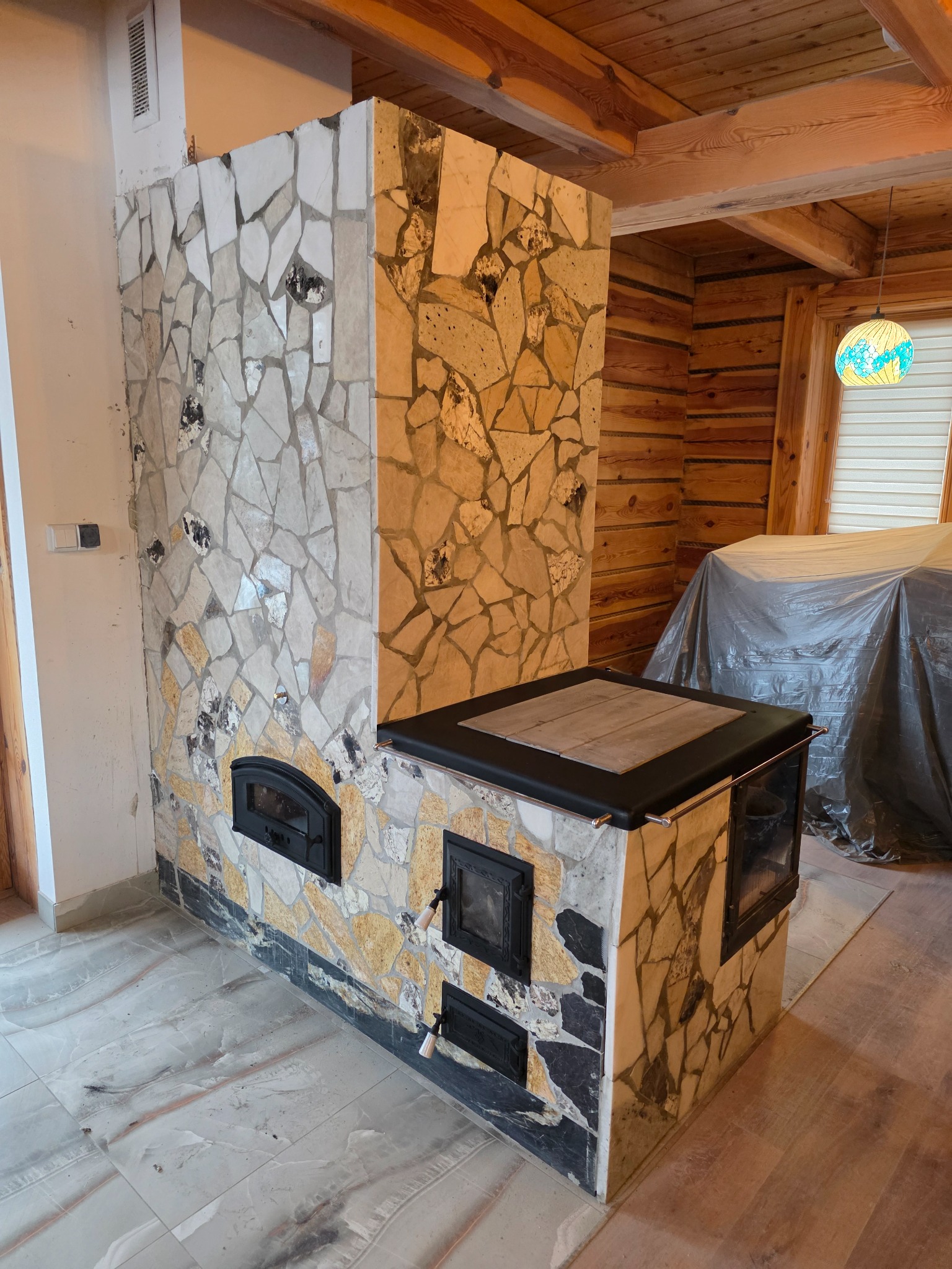 Ozdobny piec kaflowy z mozaiką z kamieni w rustykalnym wnętrzu z drewnianą boazerią. Widoczne drzwiczki paleniska i popielnika.