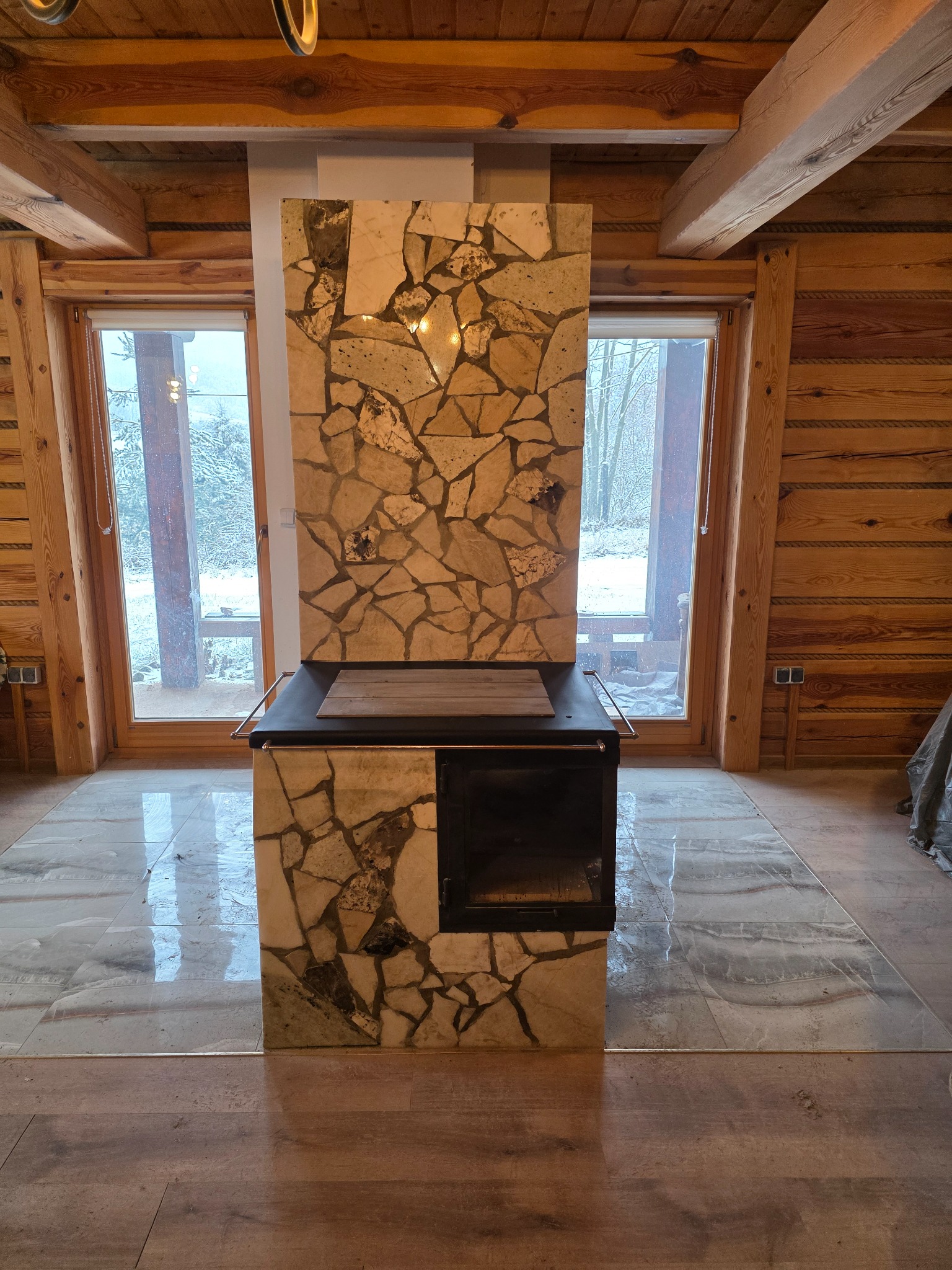 Wykończony piec kaflowy z kamienną obudową w drewnianym domu. Widoczne okna z zimowym krajobrazem. Piec z czarnymi drzwiczkami i kamienną mozaiką.