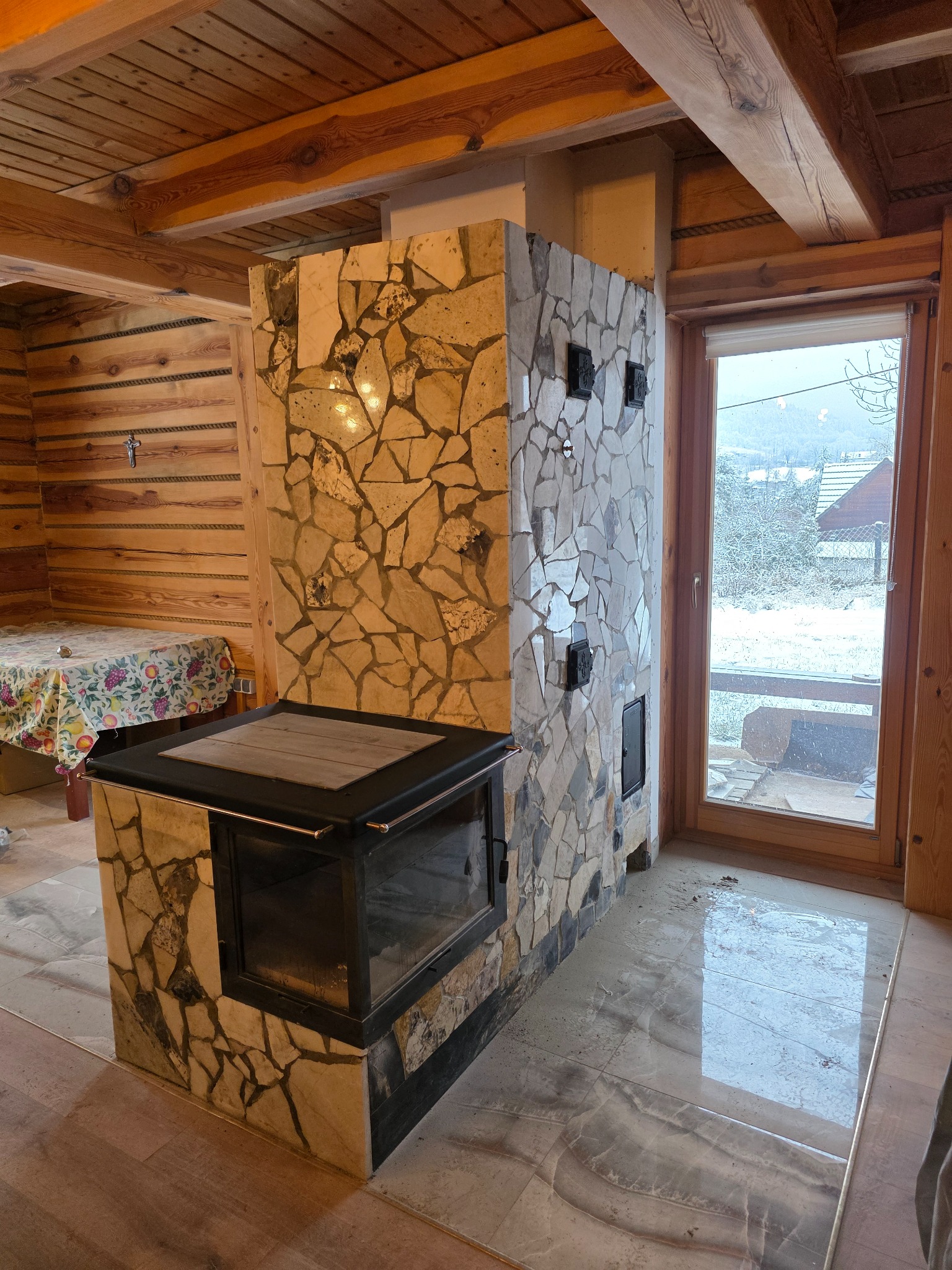 Rustykalny kominek obudowany mozaiką z kamienia w drewnianym wnętrzu. Widoczne palenisko z szybą i fragment drewnianej ściany z krzyżem. Za oknem zimowy krajobraz.