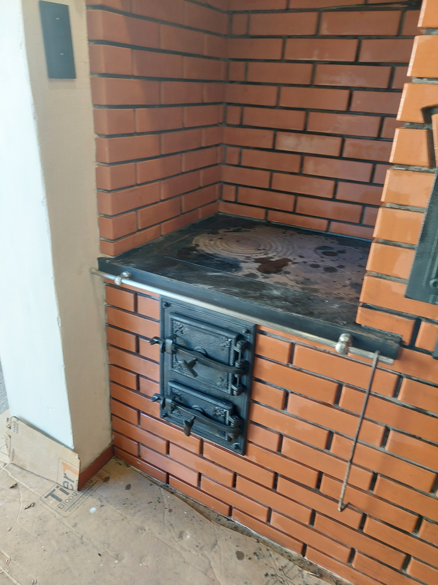 Murowana kuchnia kaflowa z czarnym blatem i żeliwnymi drzwiczkami, wykończona czerwoną cegłą. Widoczne ślady użytkowania na płycie grzewczej.