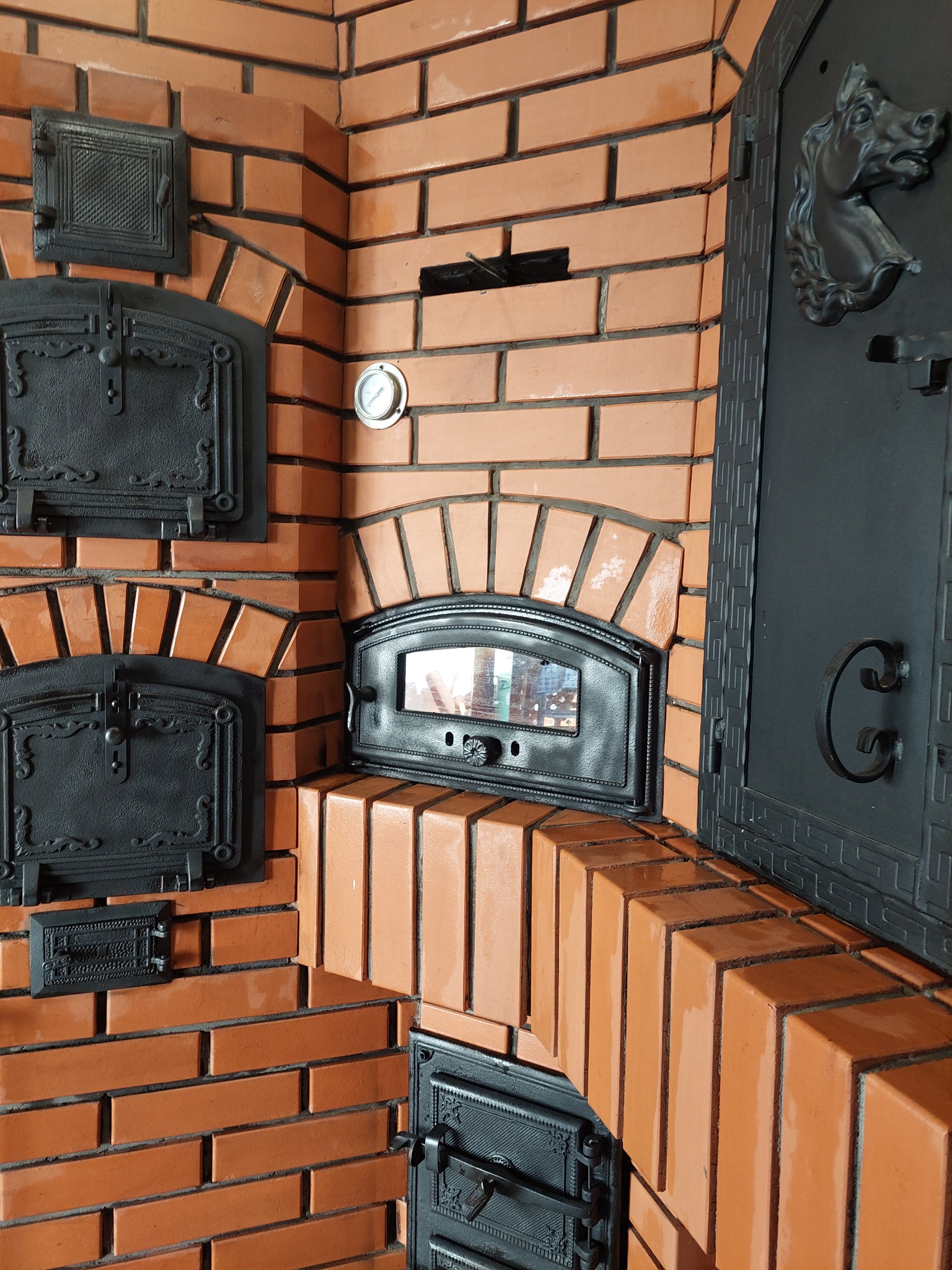 Murowany piec z cegły z czarnymi, żeliwnymi drzwiczkami i ozdobnym elementem w kształcie końskiej głowy. Widoczny termometr i detale wykończenia.