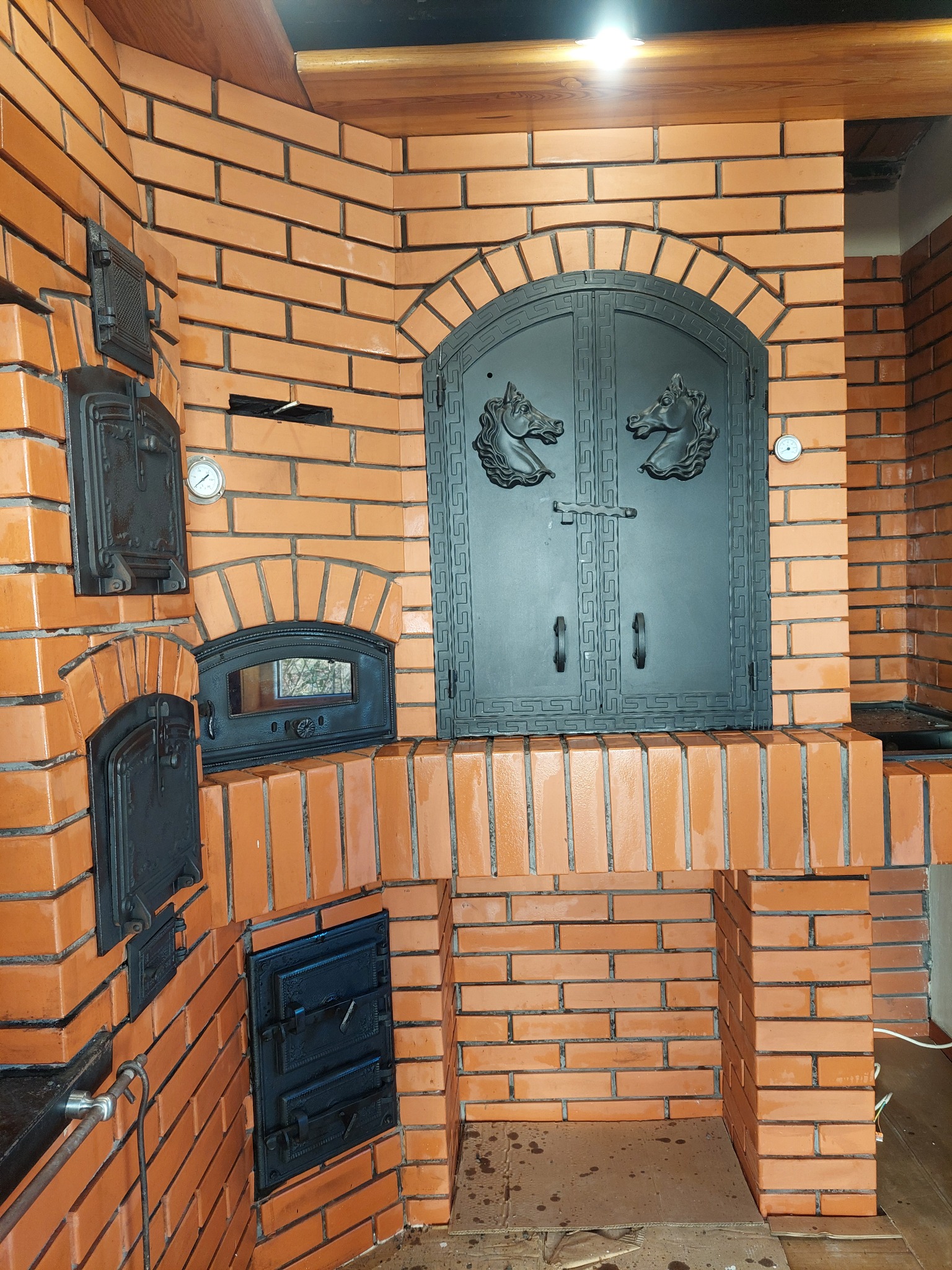 Murowany piec chlebowy z czarnymi, zdobionymi drzwiczkami i termometrami, wykończony cegłą klinkierową. Widoczne detale architektoniczne i elementy wyposażenia.