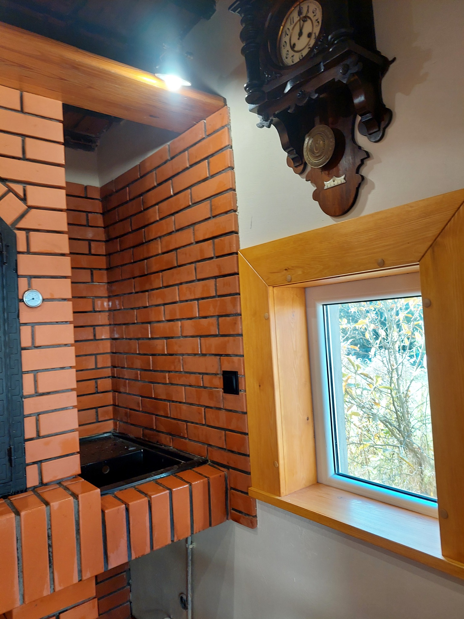 Murowany kominek z czerwonej cegły, z czarnym wkładem i termometrem, obok okno w drewnianej ramie i stary zegar na ścianie. Widok z dołu.