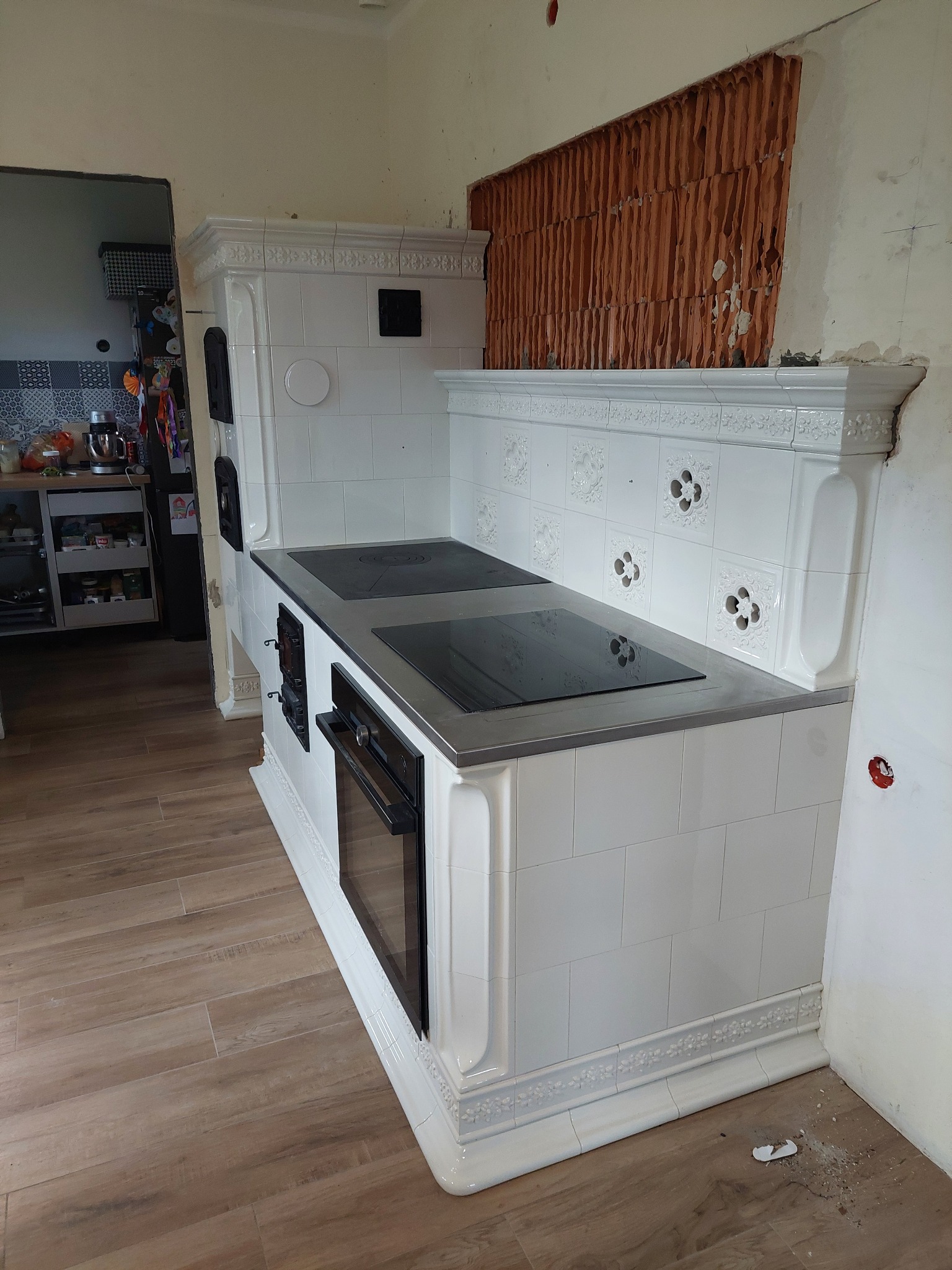 Biały, kaflowy piec kuchenny z płytą grzewczą i piekarnikiem, w trakcie montażu. Widoczne niedokończone ściany i fragment kuchni w tle. Dekoracyjne zdobienia na kaflach.