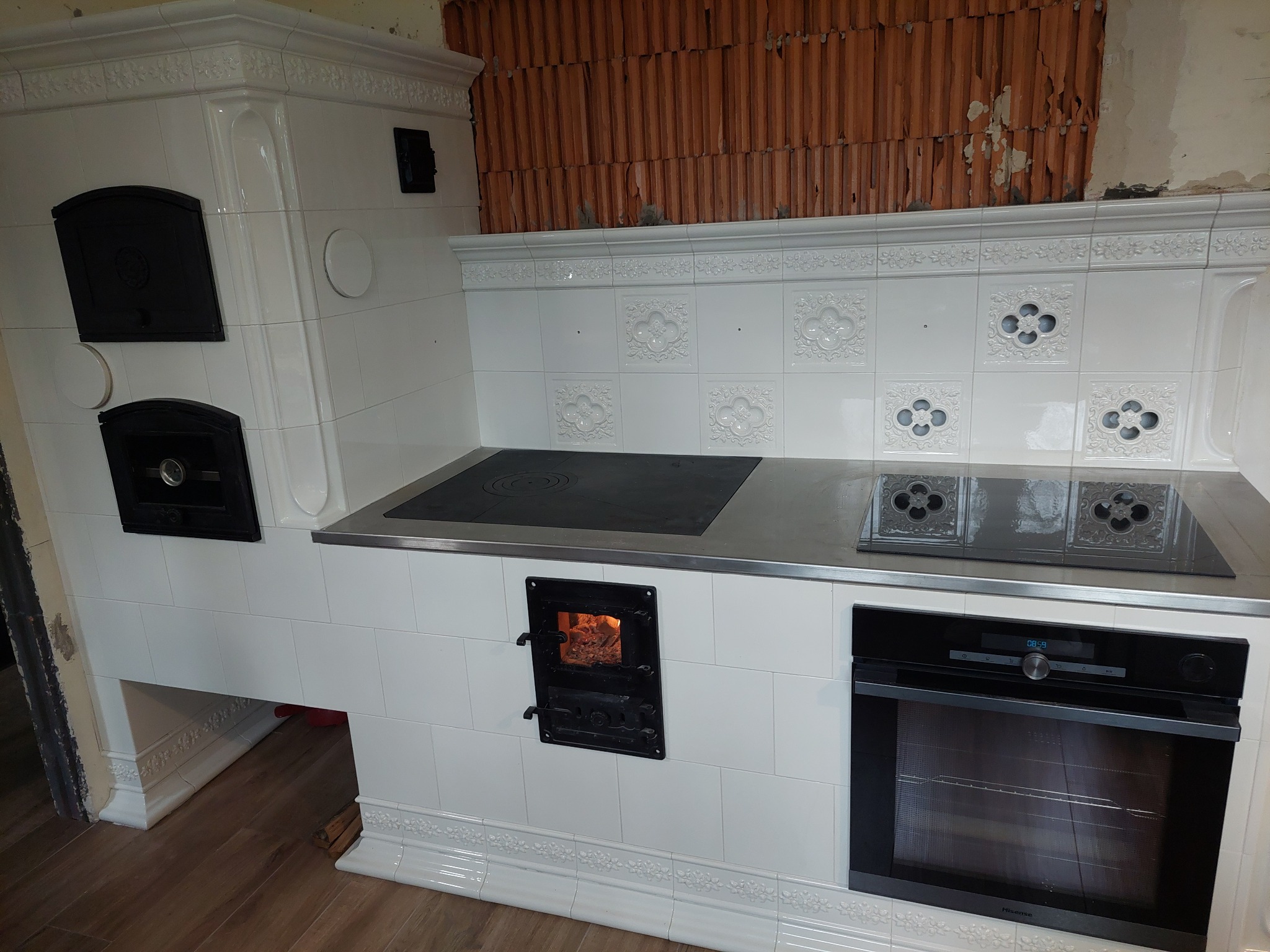 Biały, kaflowy piec kuchenny z czarnymi drzwiczkami paleniska i płytą grzewczą, wbudowany piekarnik. Widoczne cegły nad piecem. Dekoracyjne kafle.